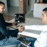 Two businessmen shake hands over a successful stock trading agreement using technology in a modern office.