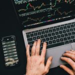Hands typing on a laptop with cryptocurrency charts and smartphone display in view, focusing on digital finance.